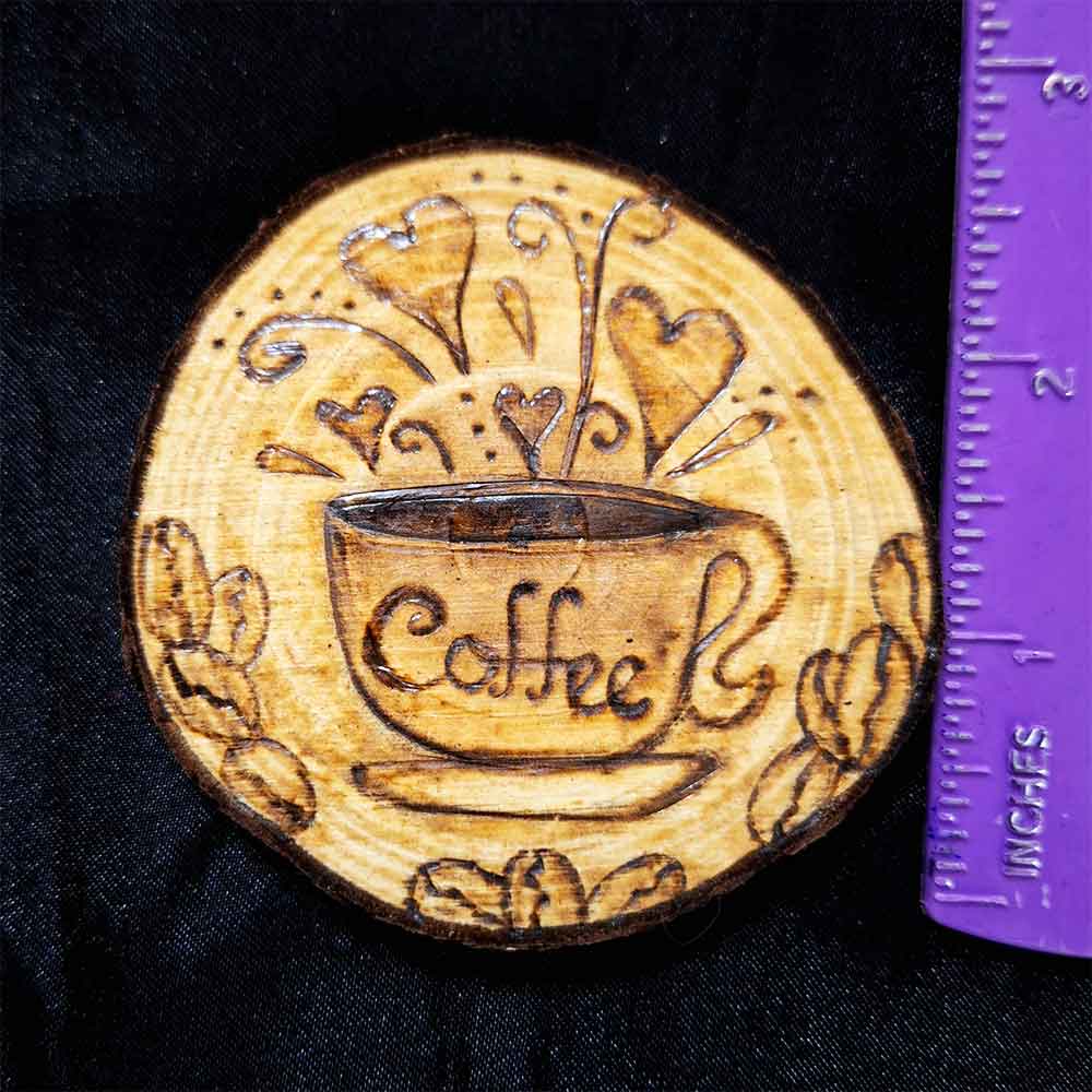 alt="Vertical ruler measuring a 3 inch by 3 inch hand burned round wood slice magnet with bark edge designed with a coffee cup on a saucer surrounded by hearts and coffee beans.">