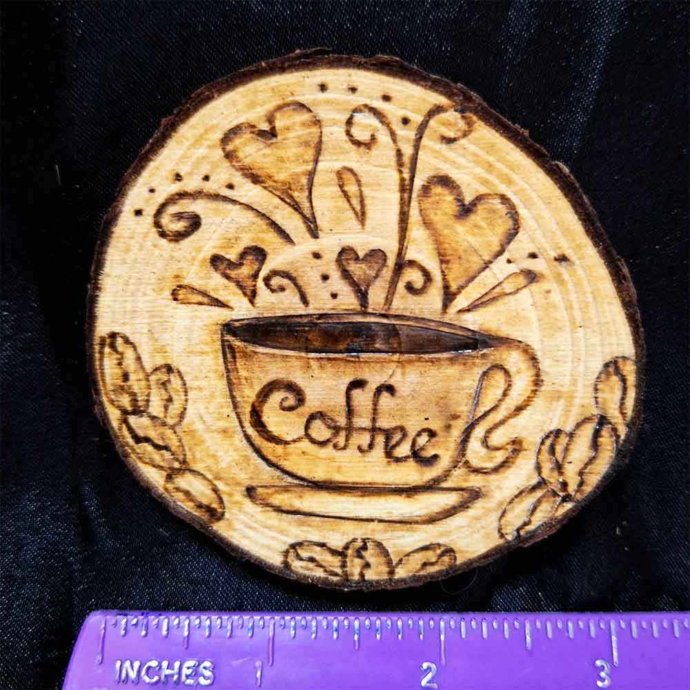alt="Horizontal ruler measuring a 3 inch by 3 inch hand burned round wood slice magnet with bark edge designed with a coffee cup on a saucer surrounded by hearts and coffee beans.">