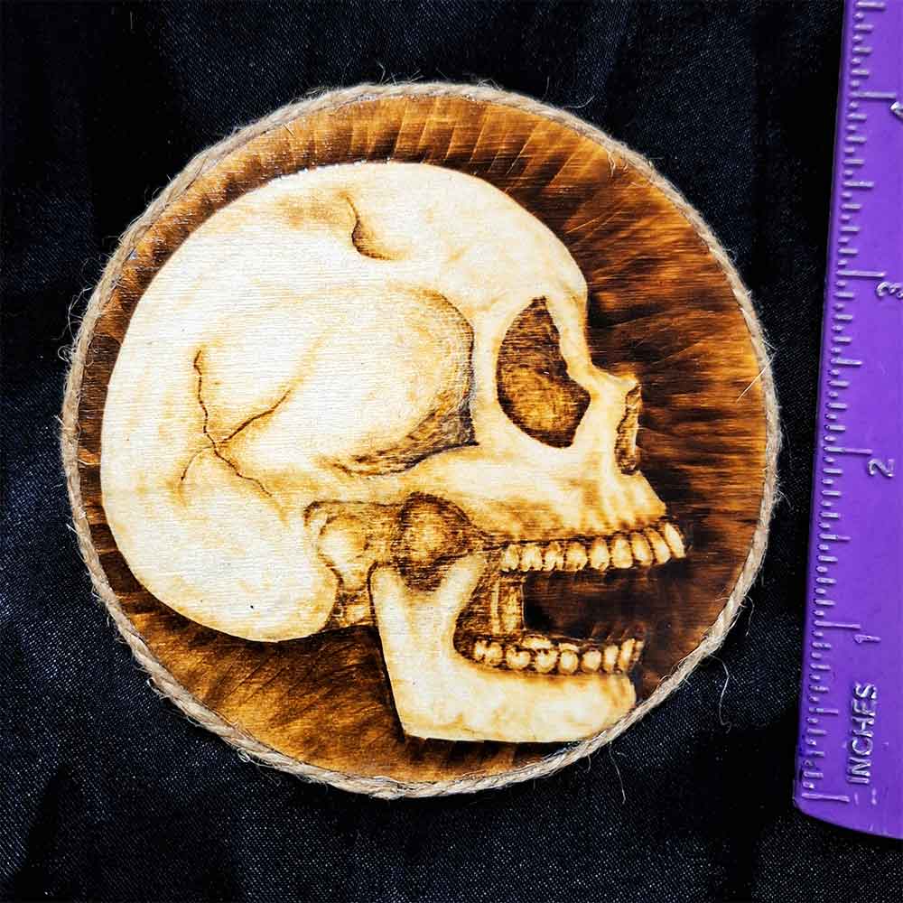 alt="Vertical ruler measuring a 4 inch by 4 inch round wood slice magnet designed with hand burned human skull art finished with a brown twine outer edge.">