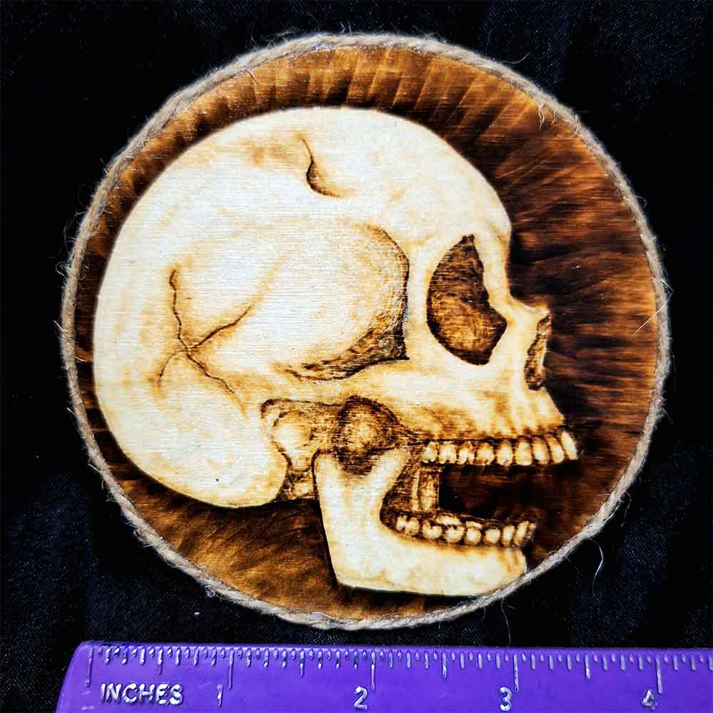 alt="Horizontal ruler measuring a 4 inch by 4 inch round wood slice magnet designed with hand burned human skull art finished with a brown twine outer edge.">