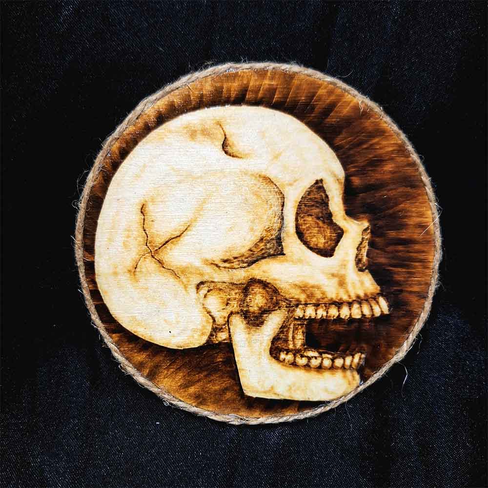 alt="4 inch by 4 inch round wood slice magnet designed with hand burned human skull art finished with a brown twine outer edge.">
