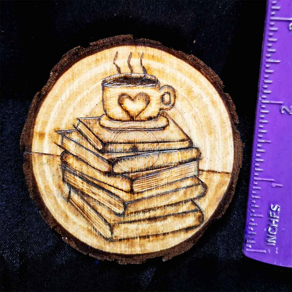alt="Vertical ruler measuring a 3 inch by 3 inch hand burned round wood slice magnet with bark edge designed with a coffee cup with a heart on a saucer sitting on top of a stack of books.">