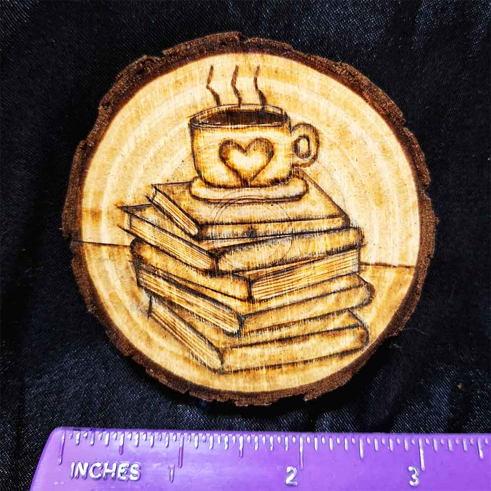 alt="Horizontal ruler measuring a 3 inch by 3 inch hand burned round wood slice magnet with bark edge designed with a coffee cup with a heart on a saucer sitting on top of a stack of books.">