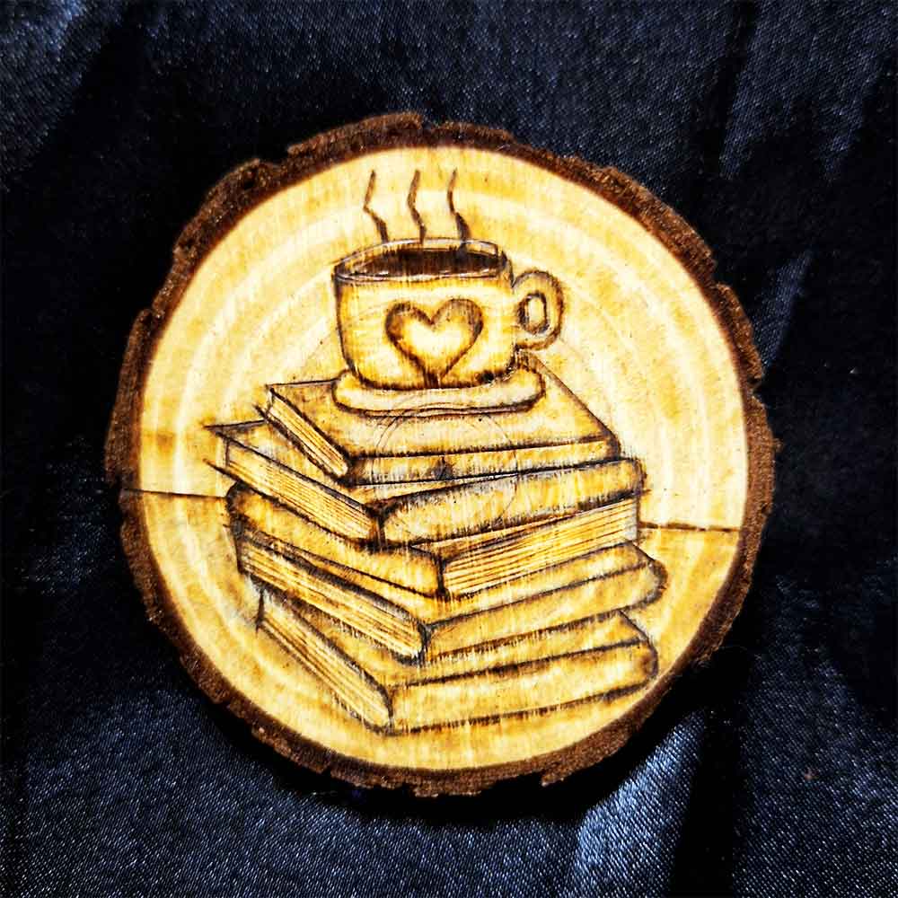 alt="3 inch by 3 inch hand burned round wood slice magnet with bark edge designed with a coffee or tea cup with a heart on a saucer sitting on top of a stack of books.">