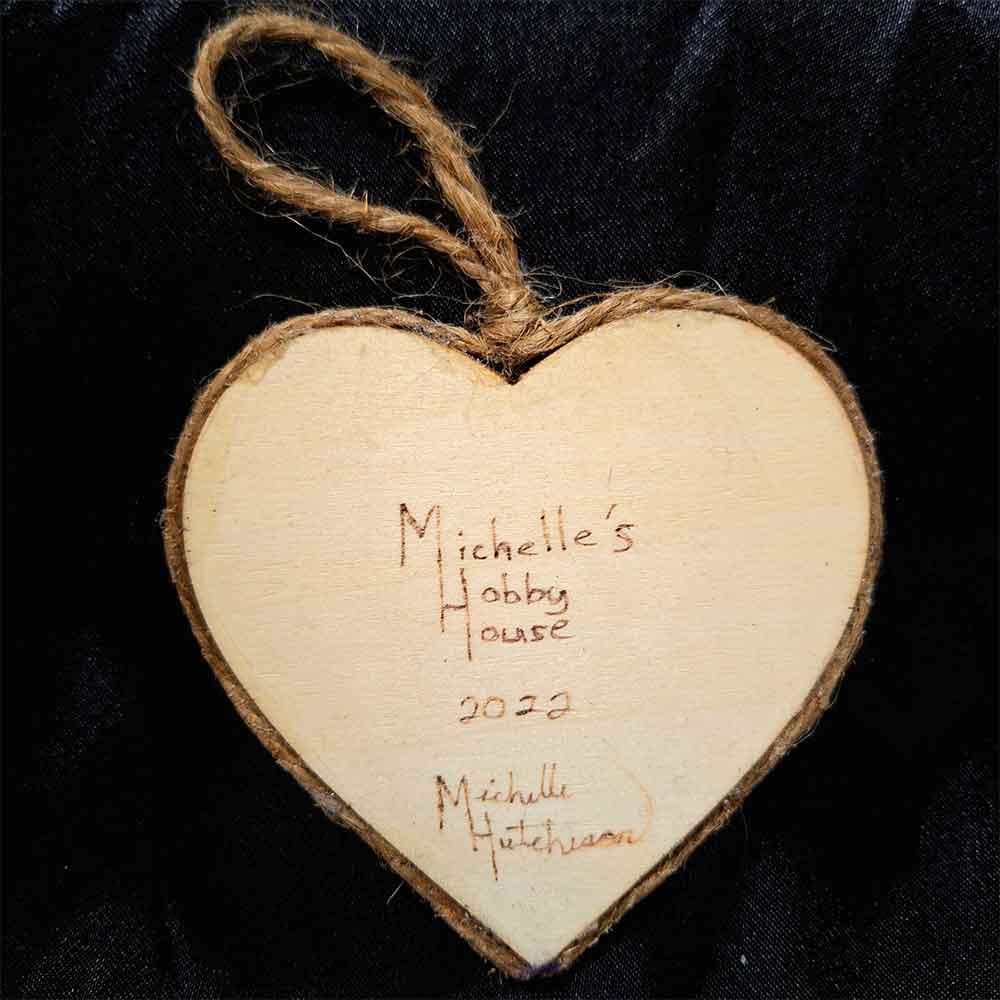 alt="View of the back artist and brand signatures on a 3 inch by 3 inch heart shaped wood slice finished with a brown twine outer edge with a loop for wall hanging.">