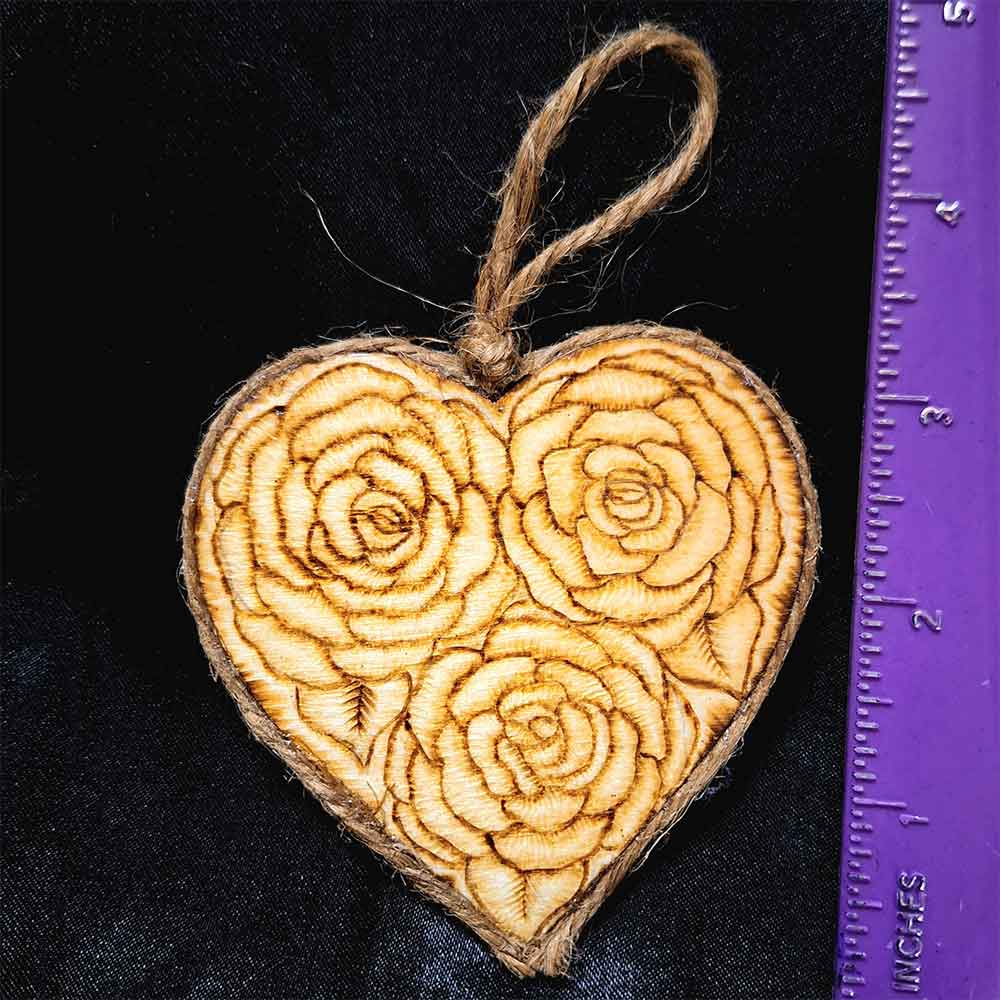 alt="Vertical ruler measuring a 3 inch by 3 inch heart shaped wood slice designed with 3 roses artwork hand burned finished with a brown twine outer edge with a loop for wall hanging.">