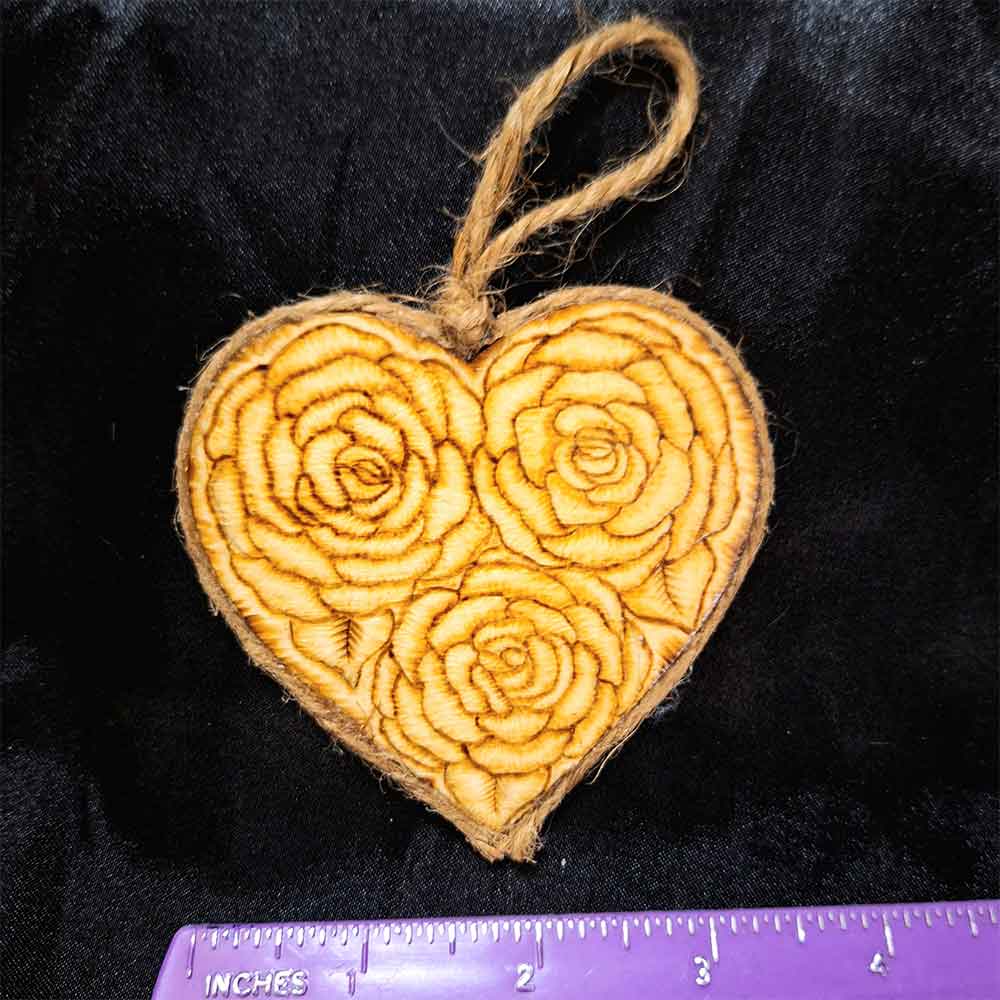 alt="3 inch by 3 inch heart shaped wood slice designed with 3 roses artwork hand burned finished with a brown twine outer edge with a loop for wall hanging.">
