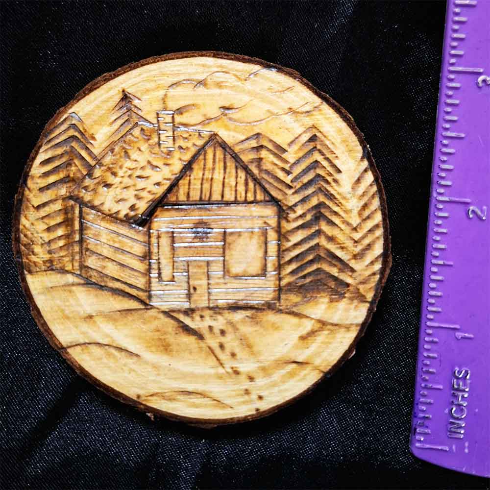 alt="Vertical ruler measuring a 3 inch by 3 inch hand burned round wood slice magnet with bark edge designed with a cozy wood cabin nestled in trees and snow with a smoking chimney and footprints in the snow up to the front door.">