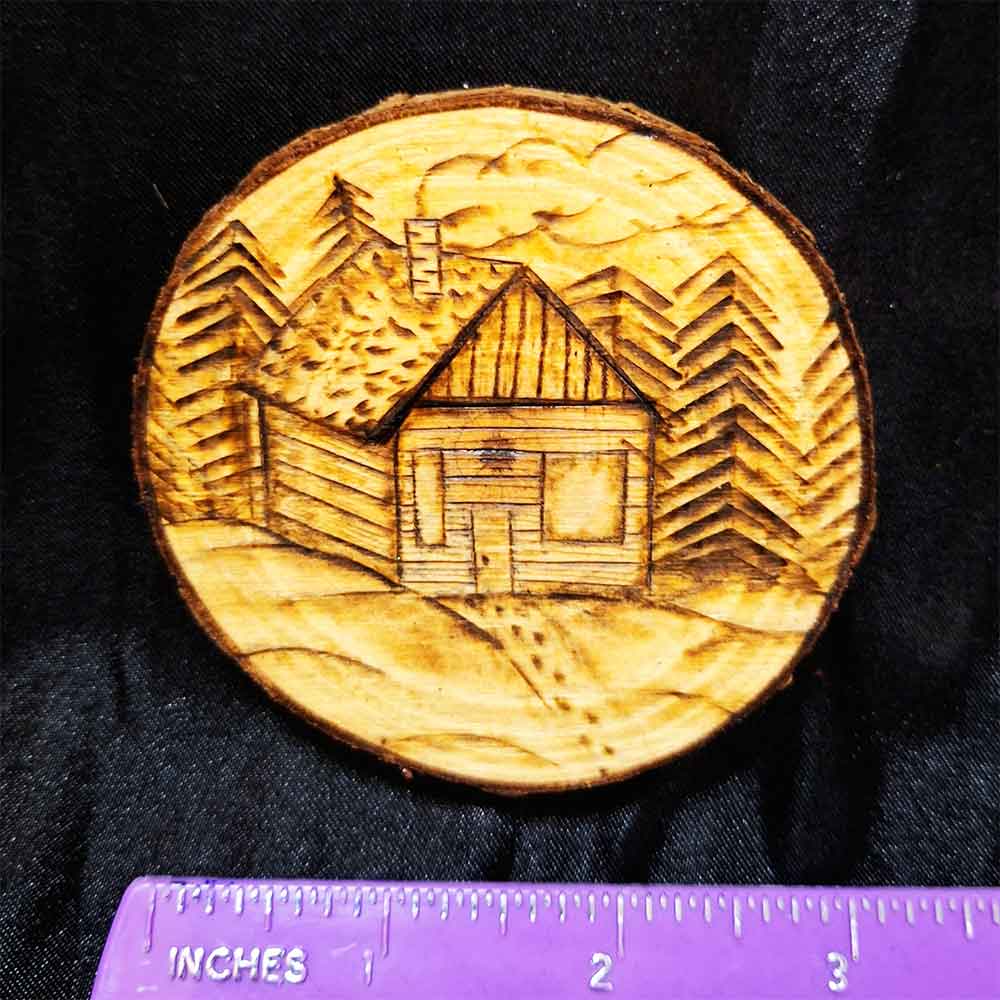 alt="Horizontal ruler measuring a 3 inch by 3 inch hand burned round wood slice magnet with bark edge designed with a cozy wood cabin nestled in trees and snow with a smoking chimney and footprints in the snow up to the front door.">