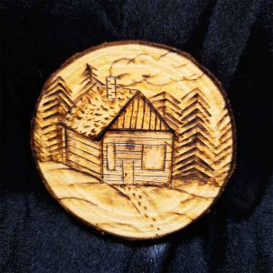 alt="3 inch by 3 inch hand burned round wood slice magnet with bark edge designed with a cozy wood cabin nestled in trees and snow with a smoking chimney and footprints in the snow up to the front door.">