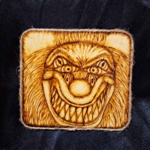 alt="4 inch by 4 inch square wood slice magnet designed with hand burned haunting three eyed clown face with a big sharp tooth smile artwork finished with a brown twine outer edge.">