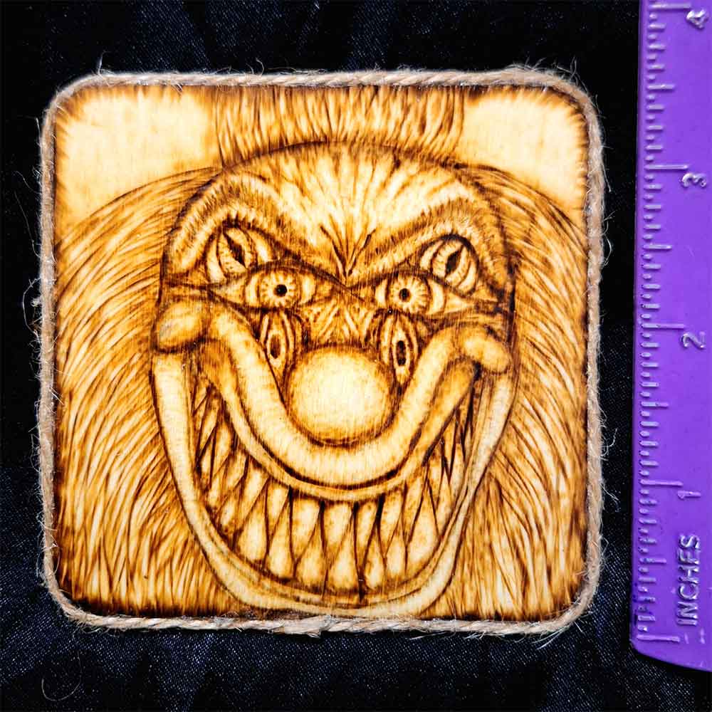 alt="Vertical ruler measuring a 4 inch by 4 inch square wood slice magnet designed with hand burned haunting three eyed clown face with a big sharp tooth smile artwork finished with a brown twine outer edge.">