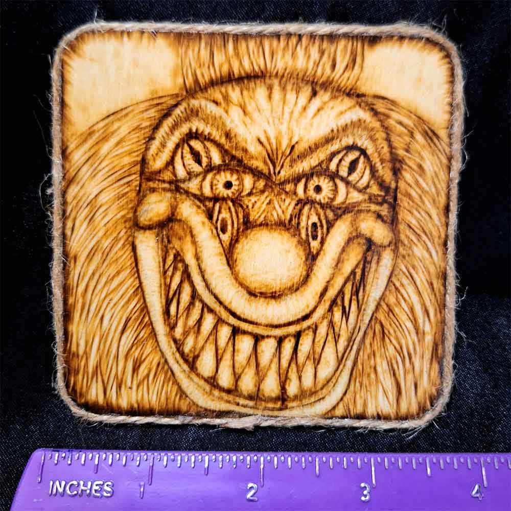 alt="Horizontal ruler measuring a 4 inch by 4 inch square wood slice magnet designed with hand burned haunting three eyed clown face with a big sharp tooth smile artwork finished with a brown twine outer edge.">