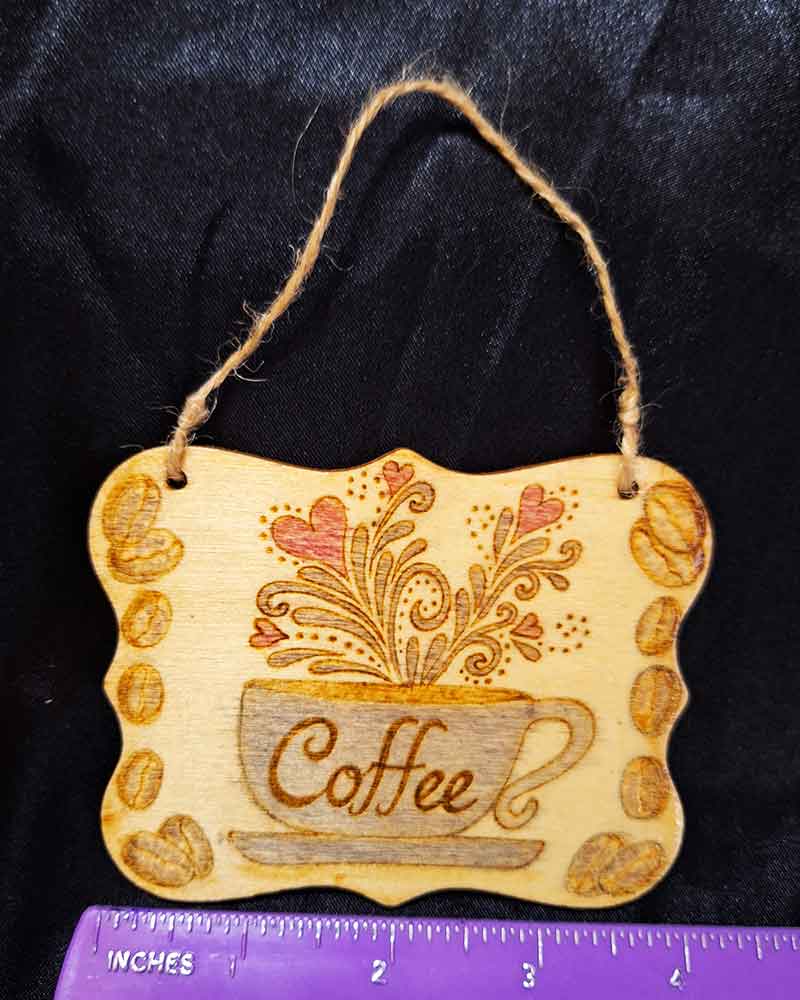 alt="Horizontal ruler measuring a 3 inch by 4 inch hand burned rectangle slice with decorative shaped edge designed with a blue coffee cup on a saucer that says 'Coffee' with red hearts and brown decorative swirl splashes coming out of the top and brown coffee beans framing the left and right sides finished with a brown twine top loop for wall hanging display.">