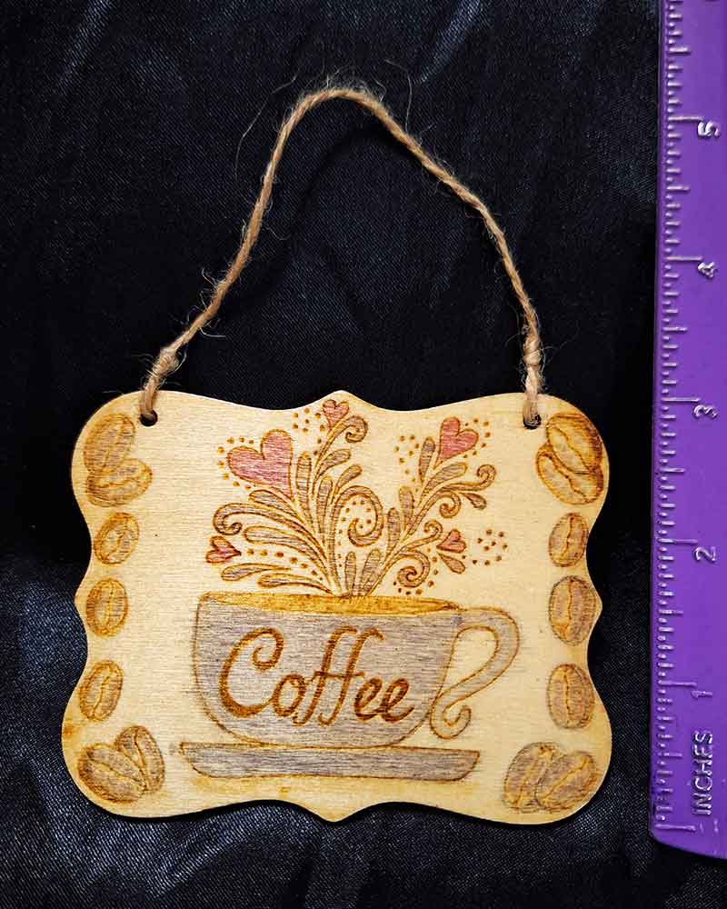 alt="Vertical ruler measuring a 3 inch by 4 inch hand burned rectangle slice with decorative shaped edge designed with a blue coffee cup on a saucer that says 'Coffee' with red hearts and brown decorative swirl splashes coming out of the top and brown coffee beans framing the left and right sides finished with a brown twine top loop for wall hanging display.">