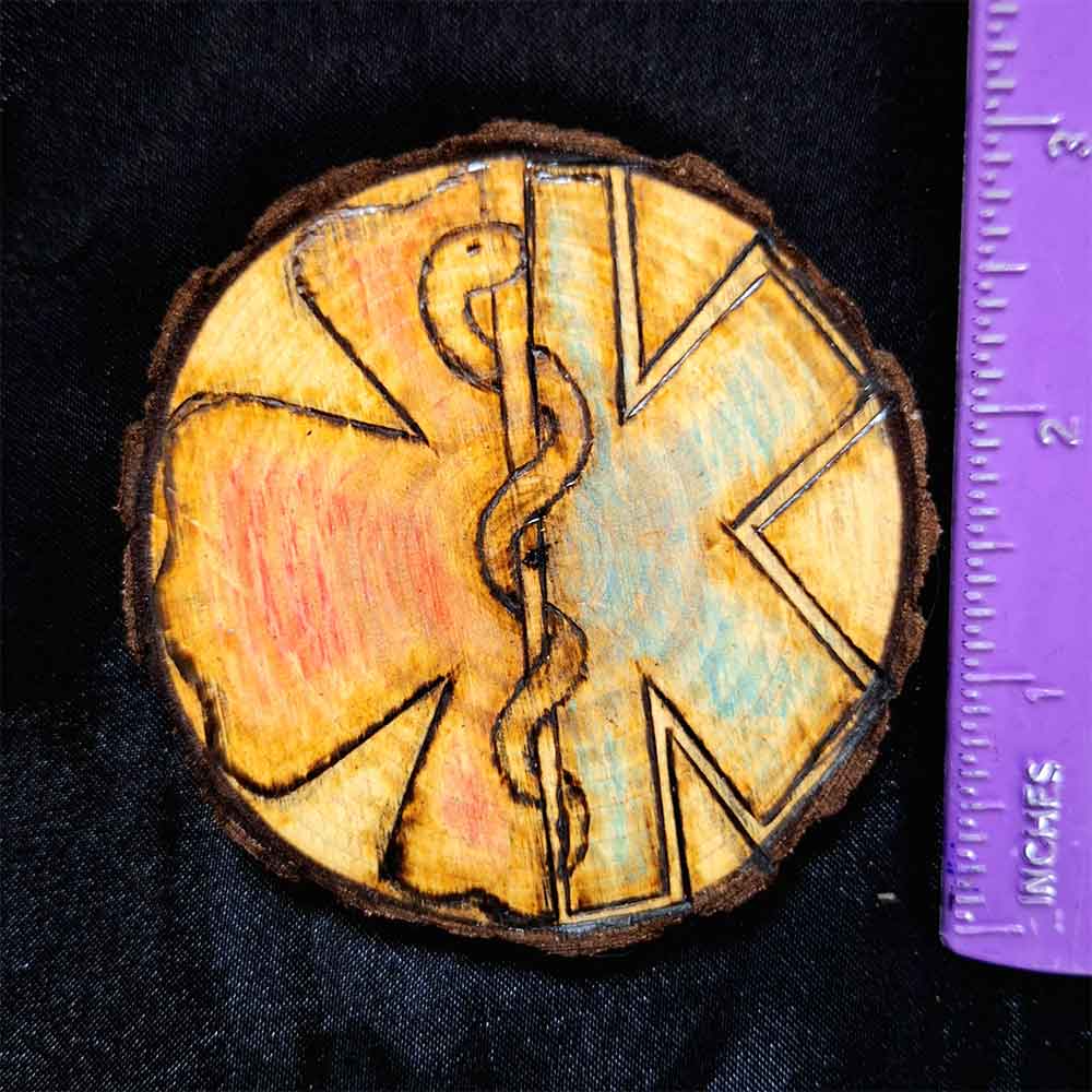 alt="Vertical ruler measuring a 3 inch by 3 inch hand burned round wood slice magnet with bark edge designed with the left half background fireman's symbol colored red and the right half background paramedics symbol colored blue with the caduceus medical symbol in the center.">
