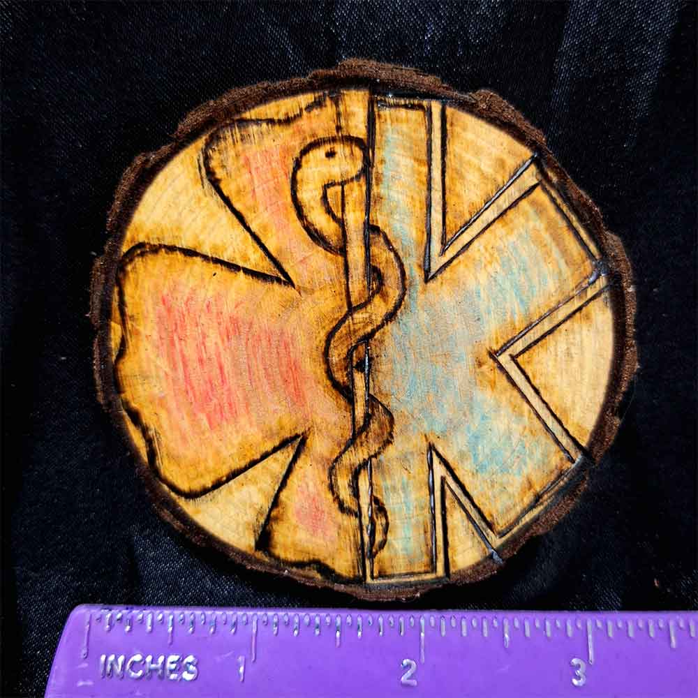 alt="Horizontal ruler measuring a 3 inch by 3 inch hand burned round wood slice magnet with bark edge designed with the left half background fireman's symbol colored red and the right half background paramedics symbol colored blue with the caduceus medical symbol in the center.">