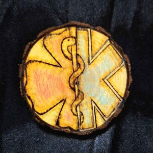 alt="3 inch by 3 inch hand burned round wood slice magnet with bark edge designed with the left half background fireman's symbol colored red and the right half background paramedics symbol colored blue with the caduceus medical symbol in the center.">