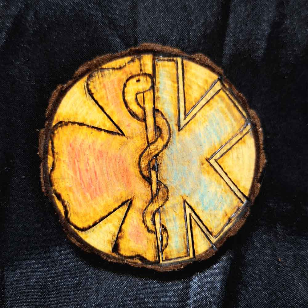 alt="3 inch by 3 inch hand burned round wood slice magnet with bark edge designed with the left half background fireman's symbol colored red and the right half background paramedics symbol colored blue with the caduceus medical symbol in the center.">