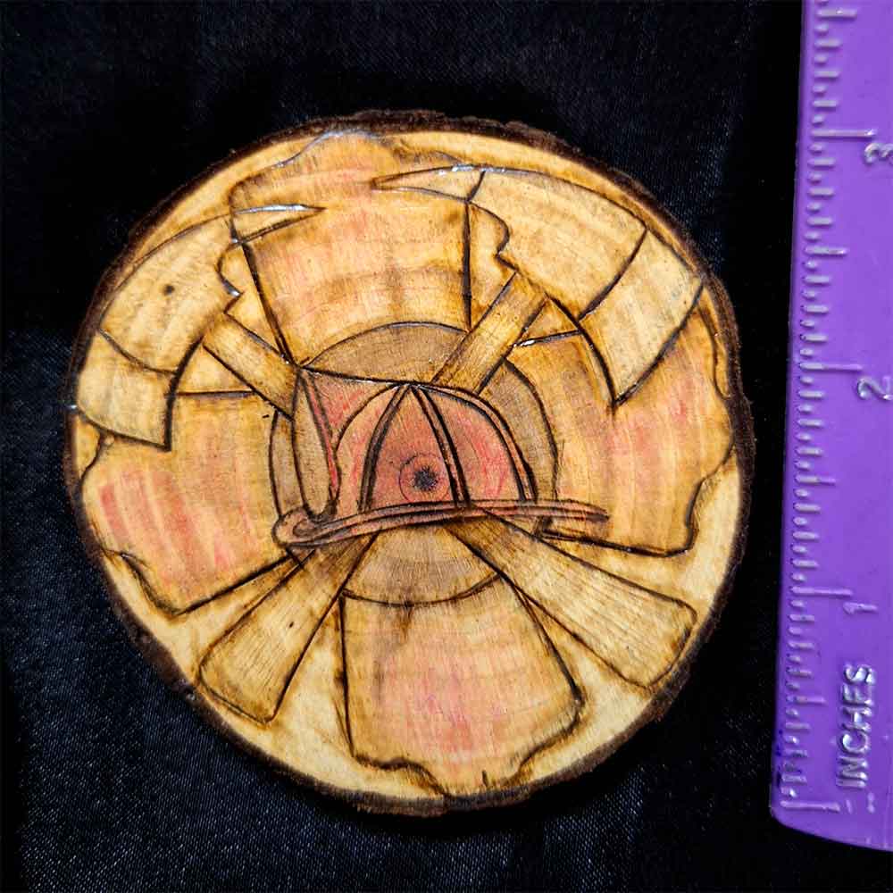 alt="Vertical ruler measuring a 3 inch by 3 inch hand burned round wood slice magnet with bark edge designed with a red colored fireman's symbol background with a red colored helmet in the center and 2 crossing axes.">