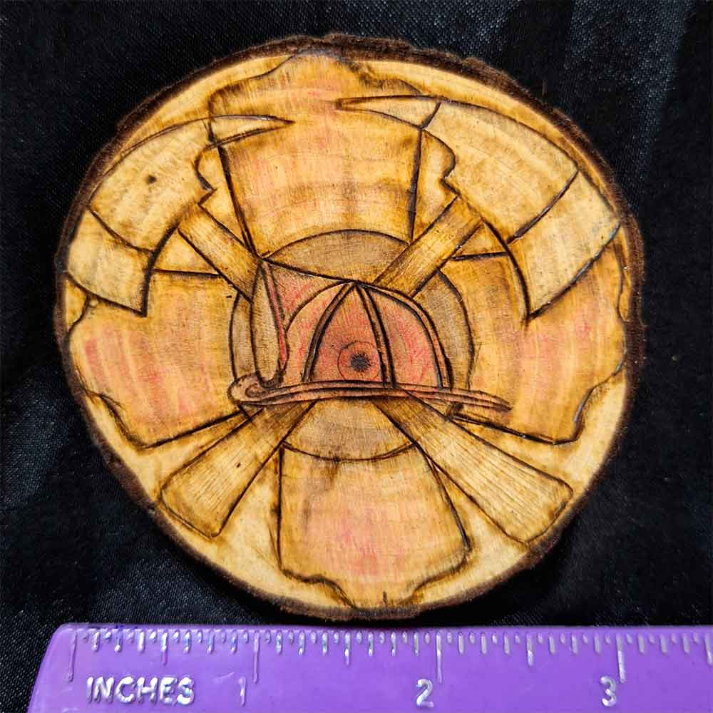 alt="Horizontal ruler measuring a 3 inch by 3 inch hand burned round wood slice magnet with bark edge designed with a red colored fireman's symbol background with a red colored helmet in the center and 2 crossing axes.">