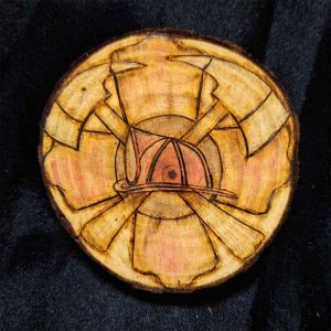 alt="3 inch by 3 inch hand burned round wood slice magnet with bark edge designed with a red colored fireman's symbol background with a red colored helmet in the center and 2 crossing axes.">