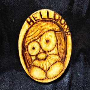 alt="2 inch by 3 inch oval wood slice magnet designed with a hand burned wide eyed gnome with a mustache peeking over his hands in a hole in a tree saying 'Hellooooo' on the border of the hole.">