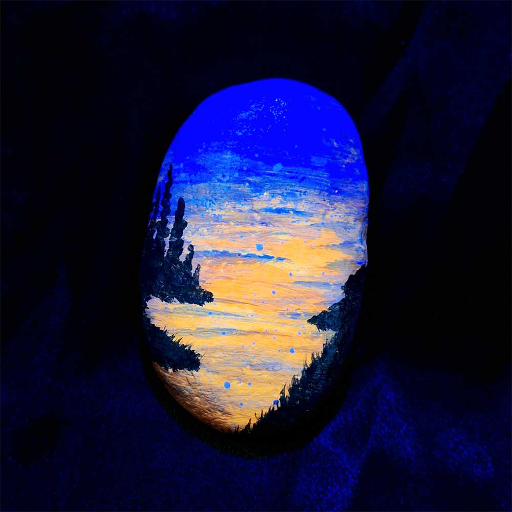 alt="Hand painted in acrylic paint 2 by 3 inch smooth oval rock with a neon orange red blue and purple blended sunset in the background with a black silhouette in the foreground containing an image of a country scene with a meandering winding river with grassy banks defining the edges and skinny pine trees in hills in the back left side of the rock viewed glowing under black light.">