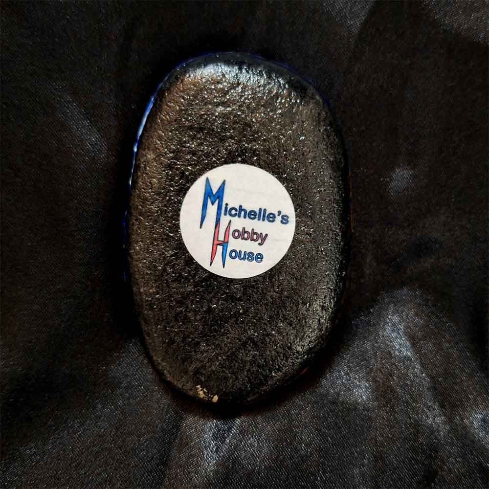 alt="View of the back artist brand label on a Hand painted 2 inch smooth oval round rock with black acrylic painted base coat.">