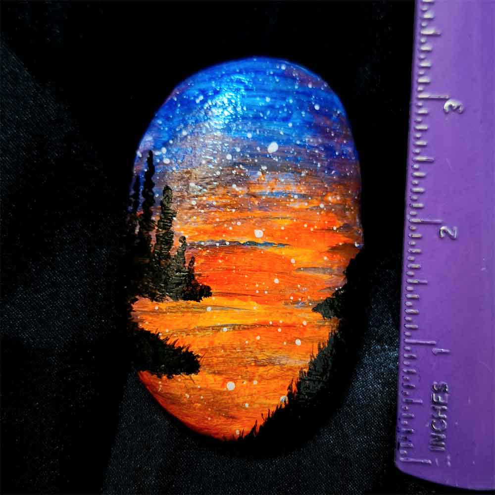 alt="Vertical ruler measuring a hand painted in acrylic paint 2 by 3 inch smooth oval rock with a neon orange red blue and purple blended sunset in the background with a black silhouette in the foreground containing an image of a country scene with a meandering winding river with grassy banks defining the edges and skinny pine trees in hills in the back left side of the rock.">