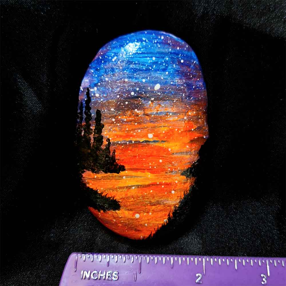 alt="Horizontal ruler measuring a hand painted in acrylic paint 2 by 3 inch smooth oval rock with a neon orange red blue and purple blended sunset in the background with a black silhouette in the foreground containing an image of a country scene with a meandering winding river with grassy banks defining the edges and skinny pine trees in hills in the back left side of the rock.">