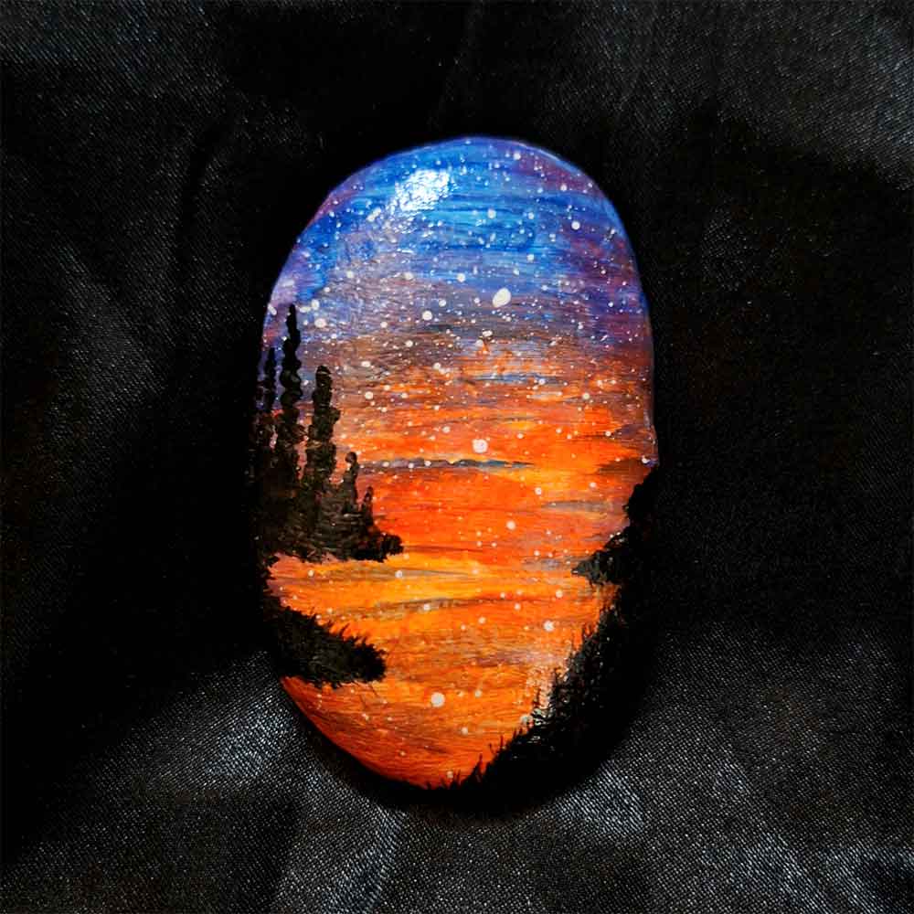 alt="Hand painted in acrylic paint 2 by 3 inch smooth oval rock with a neon orange red blue and purple blended sunset in the background with a black silhouette in the foreground containing an image of a country scene with a meandering winding river with grassy banks defining the edges and skinny pine trees in hills in the back left side of the rock.">