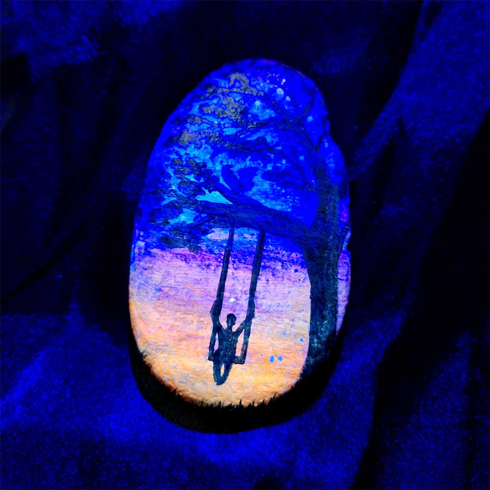 alt="Hand painted in acrylic paint 2.5 by 3 inch smooth oval rock with a neon orange, pink, purple and blue blended sunset in the background with a black silhouette in the foreground containing an image of a country scene with a tall thick branchy leafy tree on the right side and framing over the top of the rock with a person swinging on a swing hanging from a branch and grass at the bottom of the rock viewed glowing under black light.">