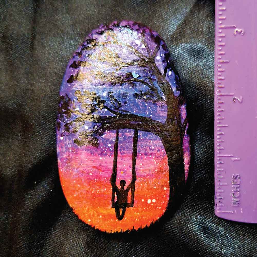 alt="Vertical ruler measuring a hand painted in acrylic paint 2.5 by 3 inch smooth oval rock with a neon orange, pink, purple and blue blended sunset in the background with a black silhouette in the foreground containing an image of a country scene with a tall thick branchy leafy tree on the right side and framing over the top of the rock with a person swinging on a swing hanging from a branch and grass at the bottom of the rock.">