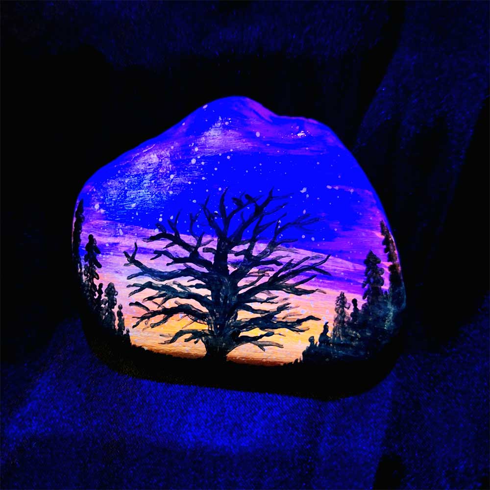 alt="Hand painted in acrylic paint 3 by 2.5 inch smooth rock with a neon orange red pink and purple blended sunset in the background with a black silhouette in the foreground containing an image of a country scene with a branchy leafless tree and grass in the center of the rock with full pine trees filling hills that are framing the far left and right sides of the rock viewed glowing under black light.">