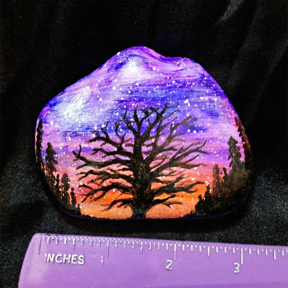 alt="Horizontal ruler measuring a hand painted in acrylic paint 3 by 2.5 inch smooth rock with a neon orange red pink and purple blended sunset in the background with a black silhouette in the foreground containing an image of a country scene with a branchy leafless tree and grass in the center of the rock with full pine trees filling hills that are framing the far left and right sides of the rock.">