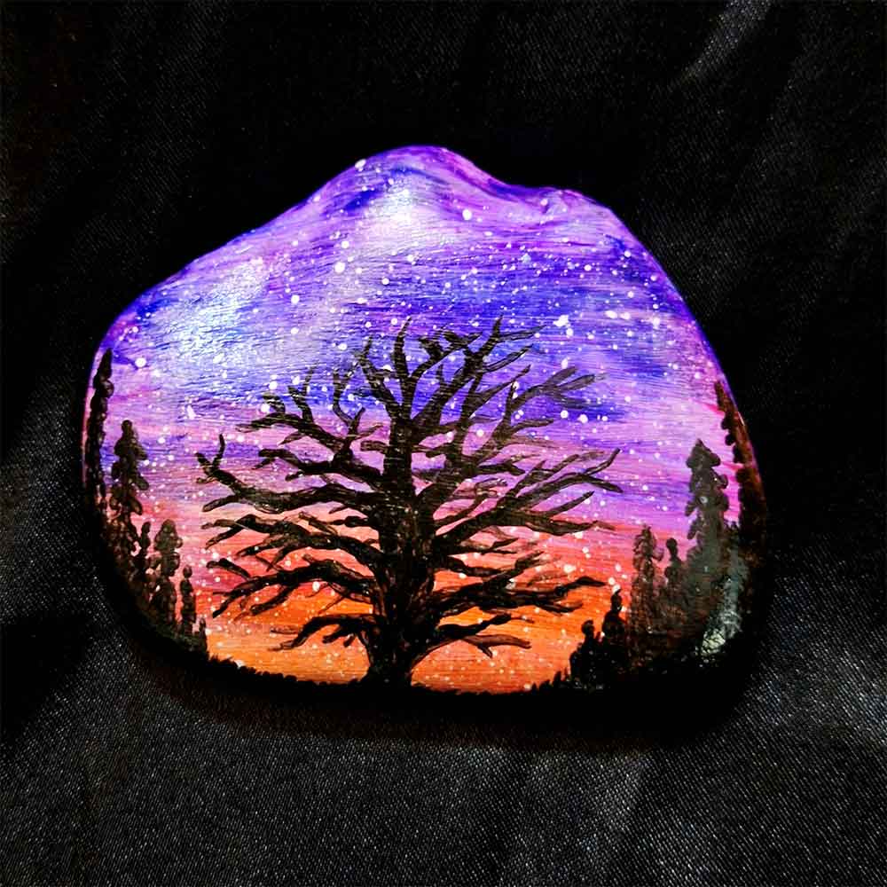 alt="Hand painted in acrylic paint 3 by 2.5 inch smooth rock with a neon orange red pink and purple blended sunset in the background with a black silhouette in the foreground containing an image of a country scene with a branchy leafless tree and grass in the center of the rock with full pine trees filling hills that are framing the far left and right sides of the rock.">