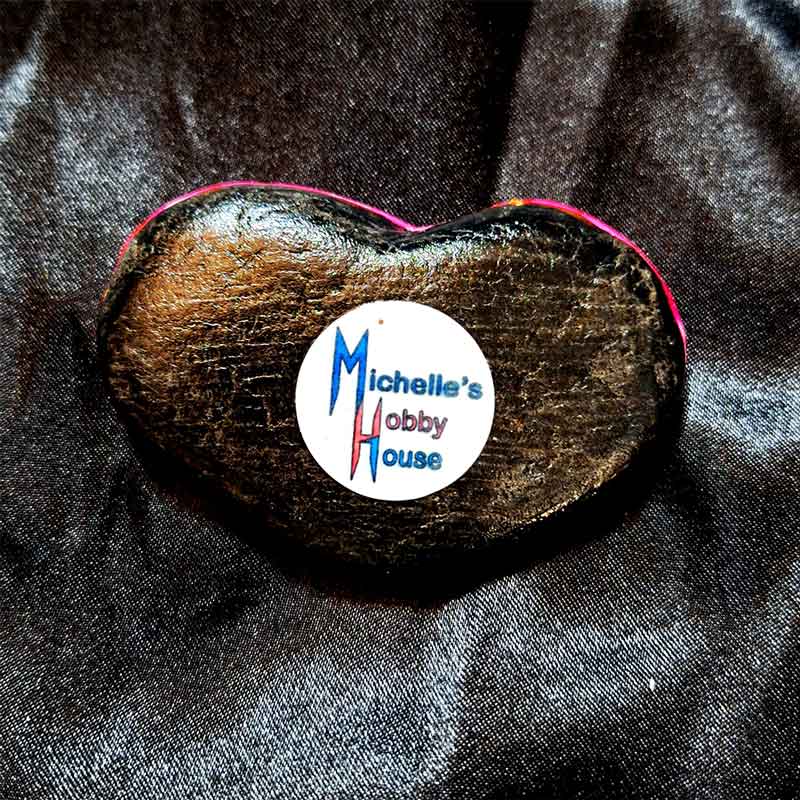 alt="View of the back artist brand label on a Hand painted 2 inch smooth round rock with black acrylic painted base coat.">