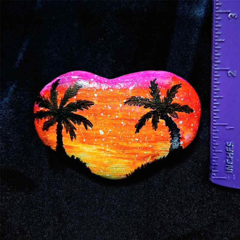alt="Vertical ruler measuring a hand painted in acrylic paint 3 by 2 inch smooth heart shaped rock with a neon yellow orange and pink blended sunset in the background with a black silhouette image of 2 tropical palm trees and grass framing the left and right sides of the rock in the foreground.">