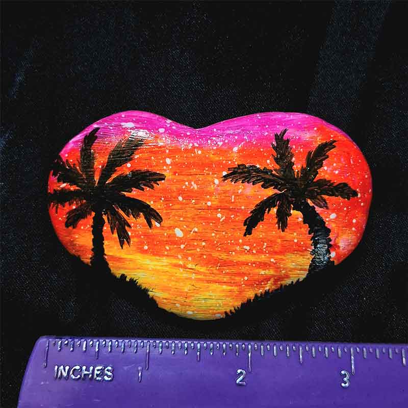 alt="Horizontal ruler measuring a hand painted in acrylic paint 3 by 2 inch smooth heart shaped rock with a neon yellow orange and pink blended sunset in the background with a black silhouette image of 2 tropical palm trees and grass framing the left and right sides of the rock in the foreground.">