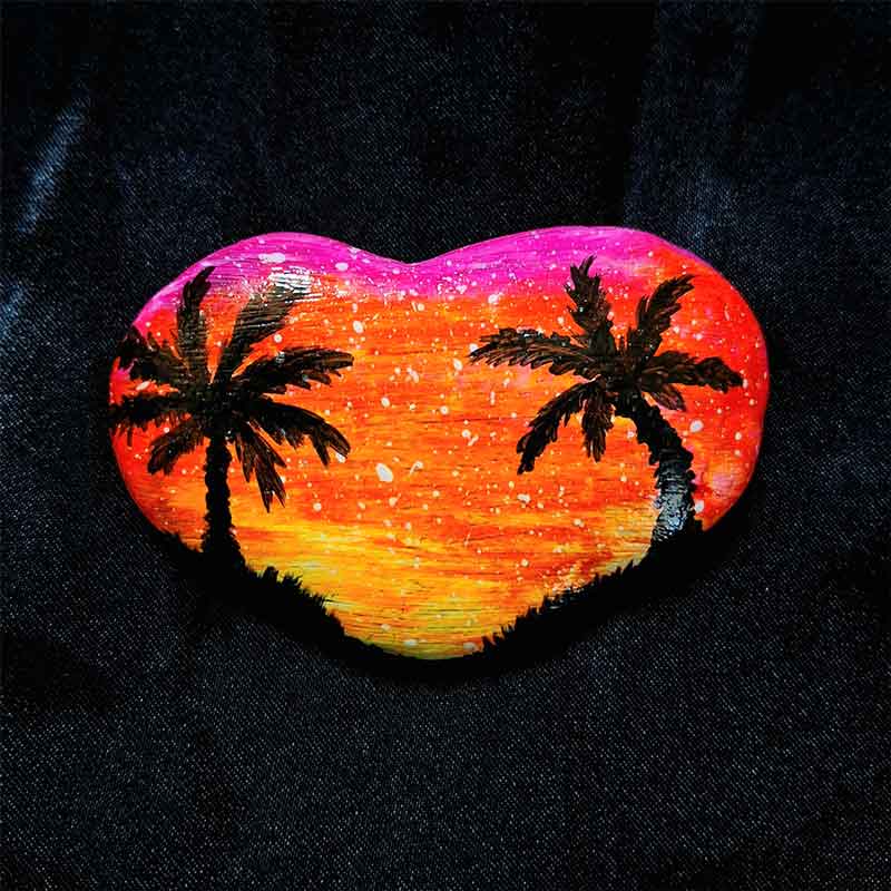 alt="Hand painted in acrylic paint 3 by 2 inch smooth heart shaped rock with a neon yellow orange and pink blended sunset in the background with a black silhouette image of 2 tropical palm trees and grass framing the left and right sides of the rock in the foreground.">