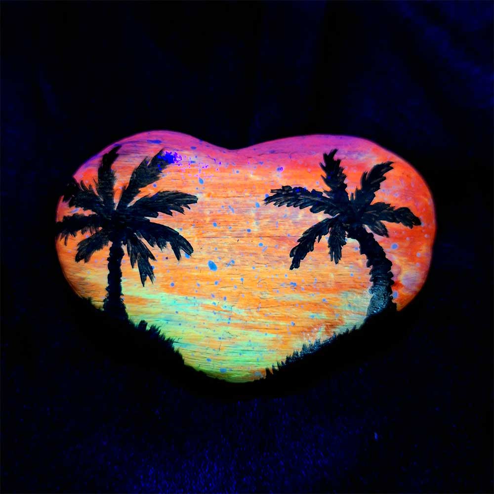 alt="Hand painted in acrylic paint 3 by 2 inch smooth heart shaped rock with a neon yellow orange and pink blended sunset in the background with a black silhouette image of 2 tropical palm trees and grass framing the left and right sides of the rock in the foreground viewed glowing under black light.">