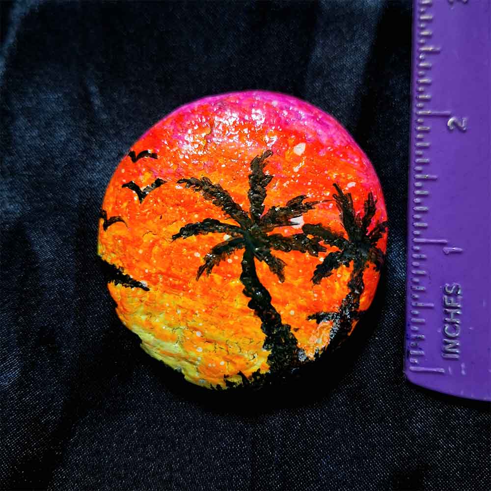 alt="Vertical ruler measuring a Hand painted in acrylic paint 2 inch smooth round rock with a neon yellow orange and pink blended sunset in the background with a black silhouette image of 2 tropical palm trees grass and birds in the foreground.">