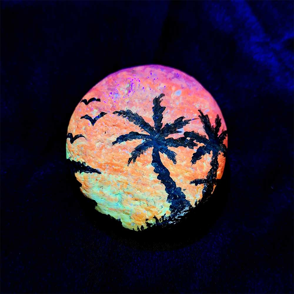 alt="View of a hand painted in acrylic paint 2 inch smooth round rock with a neon yellow orange and pink blended sunset in the background with a black silhouette image of 2 tropical palm trees grass and birds in the foreground viewed glowing under black light.">