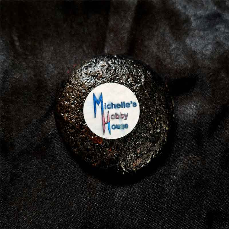 alt="View of the back artist brand label on a Hand painted 2 inch smooth round rock with black acrylic painted base coat.">