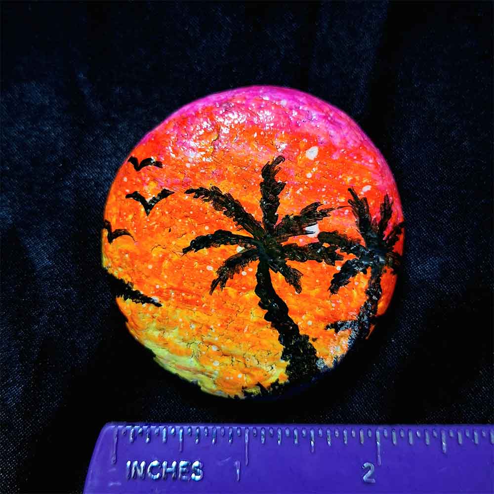 alt="Horizontal ruler measuring a Hand painted in acrylic paint 2 inch smooth round rock with a neon yellow orange and pink blended sunset in the background with a black silhouette image of 2 tropical palm trees grass and birds in the foreground.">