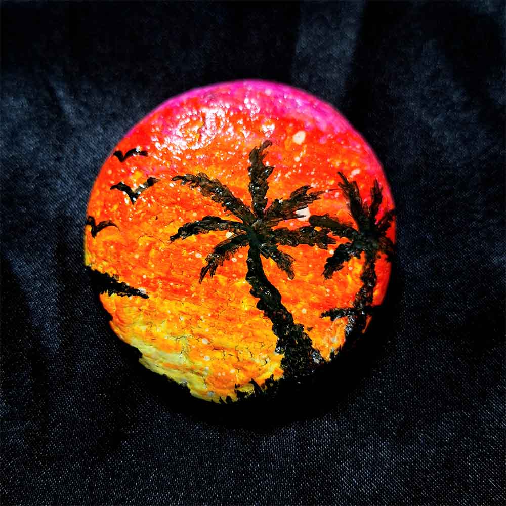 alt="Hand painted in acrylic paint 2 inch smooth round rock with a neon yellow orange and pink blended sunset in the background with a black silhouette image of 2 tropical palm trees grass and birds in the foreground.">
