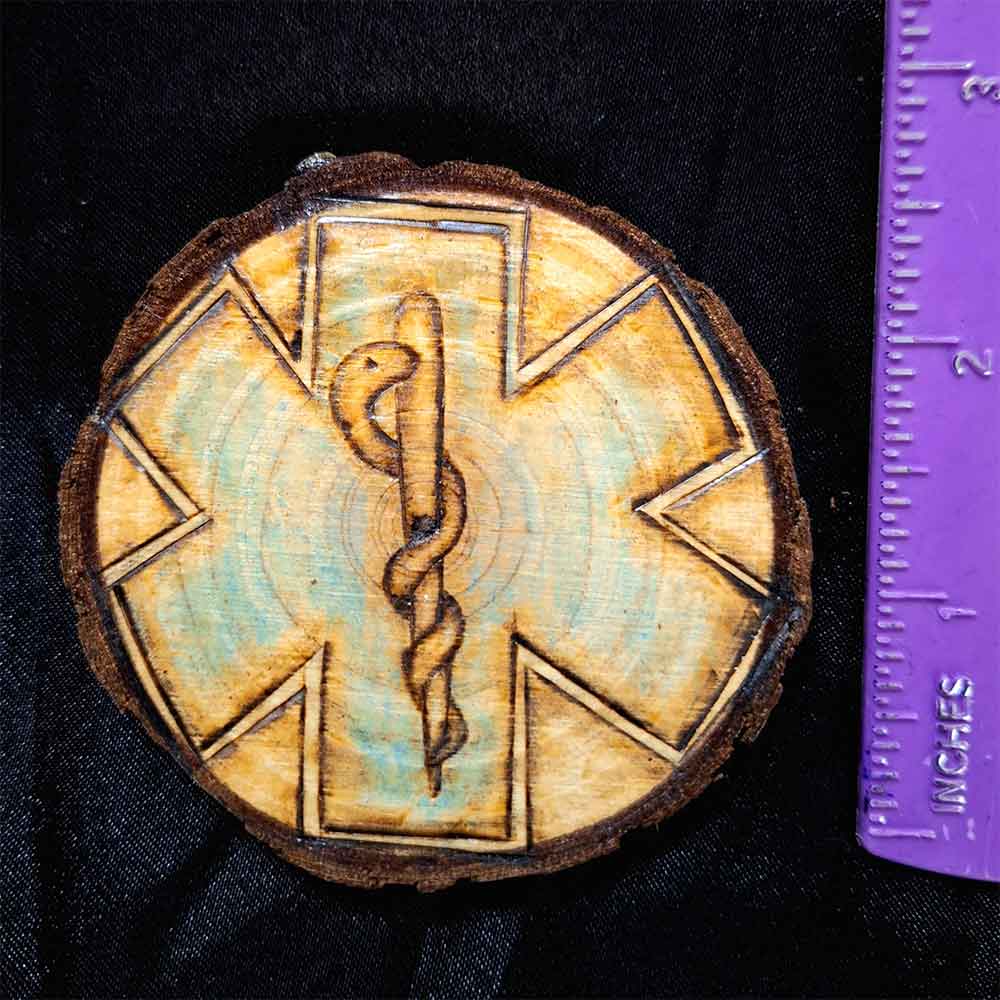 alt="Vertical ruler measuring a 3 inch by 3 inch hand burned round wood slice magnet with bark edge designed with background paramedics symbol colored blue with the caduceus medical symbol in the center.">