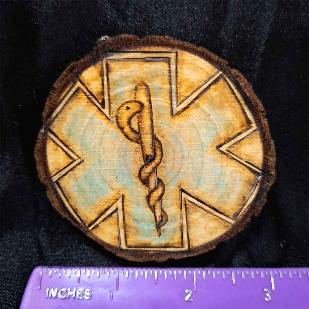 alt="Horizontal ruler measuring a 3 inch by 3 inch hand burned round wood slice magnet with bark edge designed with background paramedics symbol colored blue with the caduceus medical symbol in the center.">
