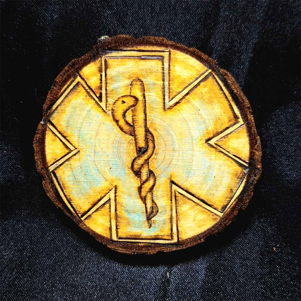 alt="3 inch by 3 inch hand burned round wood slice magnet with bark edge designed with background paramedics symbol colored blue with the caduceus medical symbol in the center.">